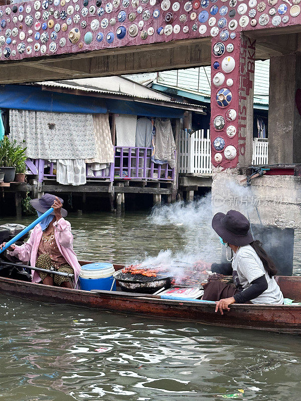 在木船上，人们在鼓式烤架上烧烤肉，游览运河附近的Khlong Damnoen Saduak，水路连接着Tha Chin和Mae Klong河